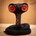 Indlæs billede i Gallery viewer, MM Figur Sacky Serpent (13 cm) er et sammenrullet, sort plysdyr med røde knapøjne og syede "X"er, perfekt som samlerfigur eller unik pynt til hylden, vist på en træbaggrund.