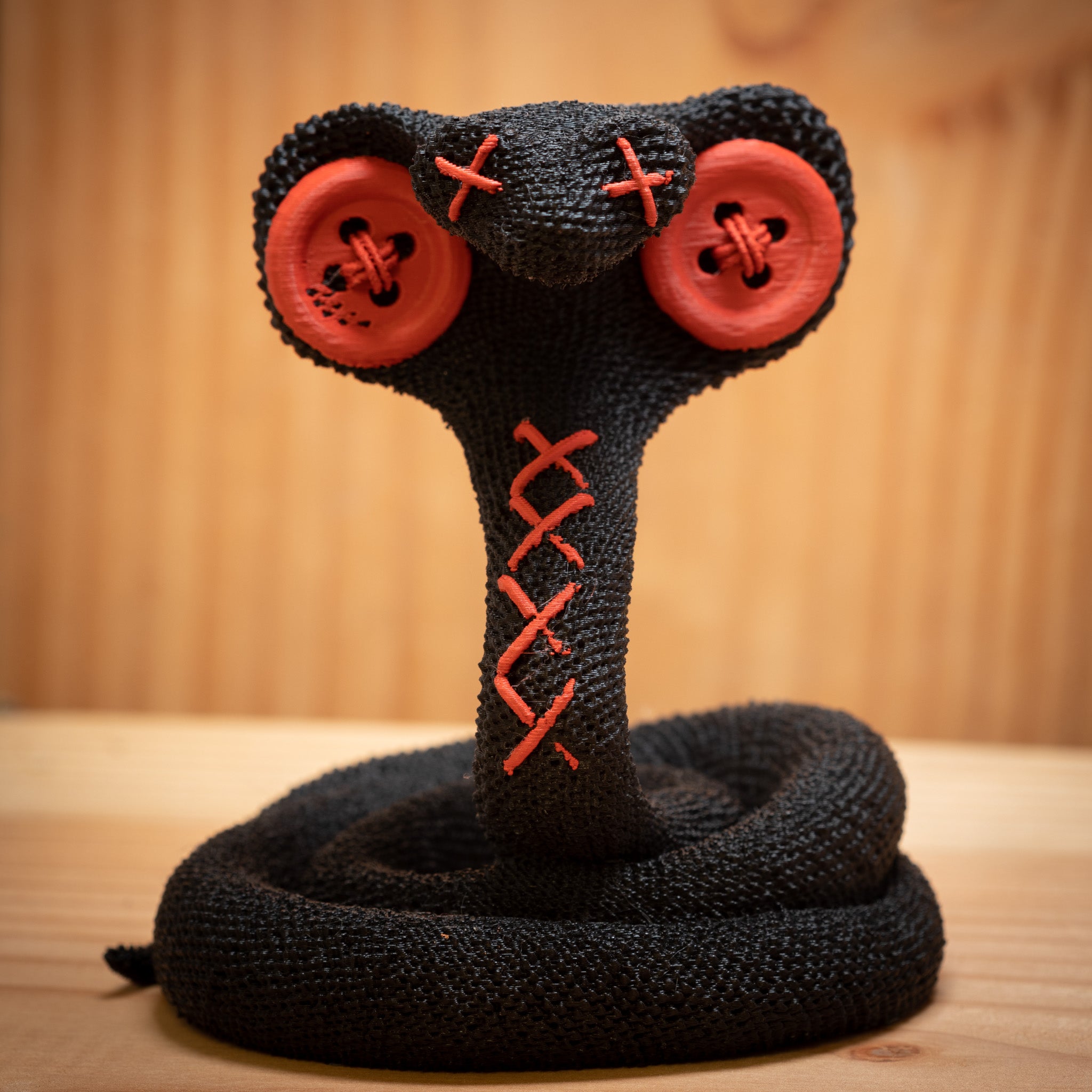 MM Figur Sacky Serpent (13 cm) er et sammenrullet, sort plysdyr med røde knapøjne og syede "X"er, perfekt som samlerfigur eller unik pynt til hylden, vist på en træbaggrund.