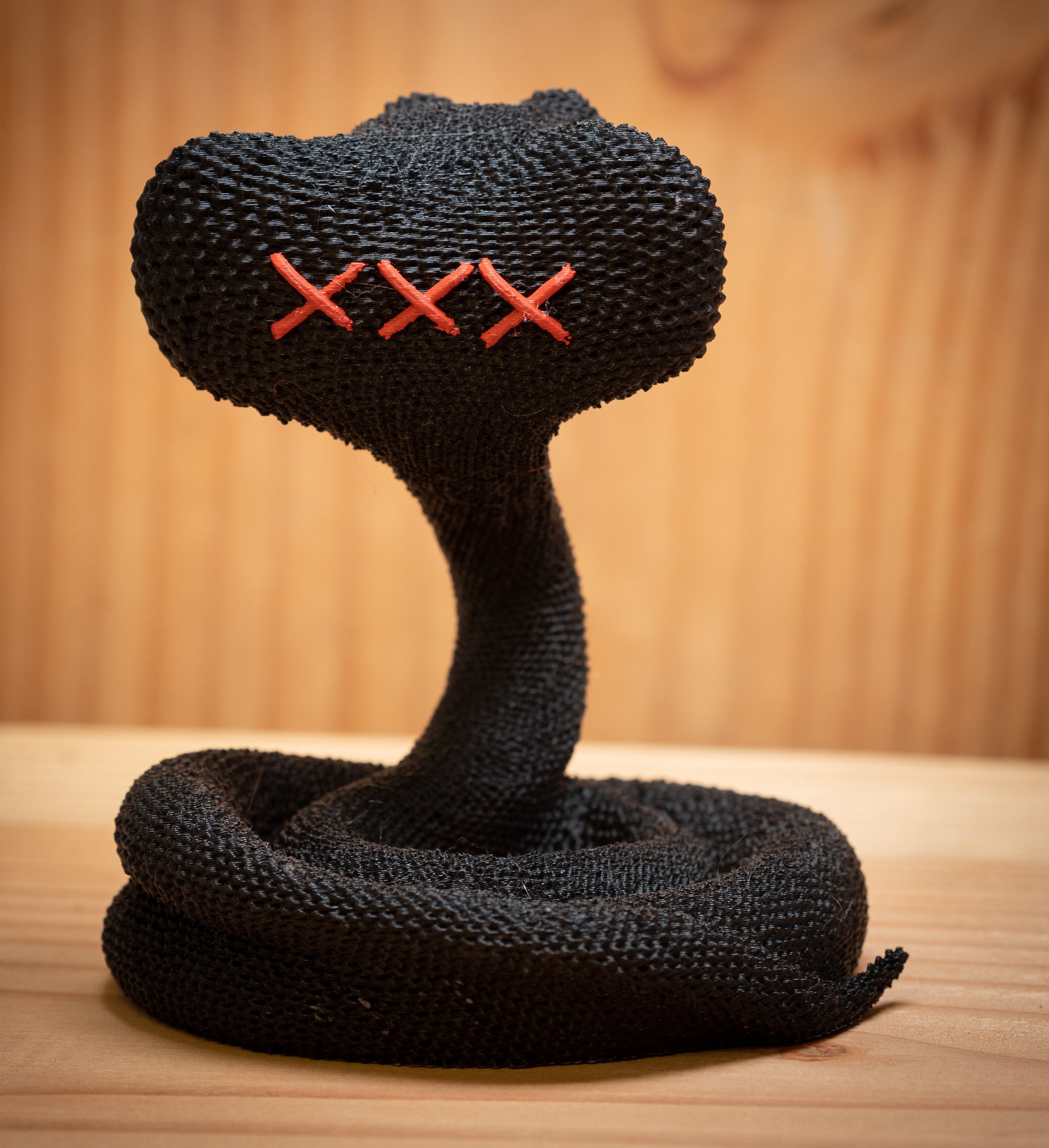 MM Figur Sacky Serpent (13 cm) er en sammenrullet sort strikket figur med tre røde syede X-øjne, der vises på en træoverflade som en iøjnefaldende samlerfigur og pynt til hylden mod en sløret træbaggrund.