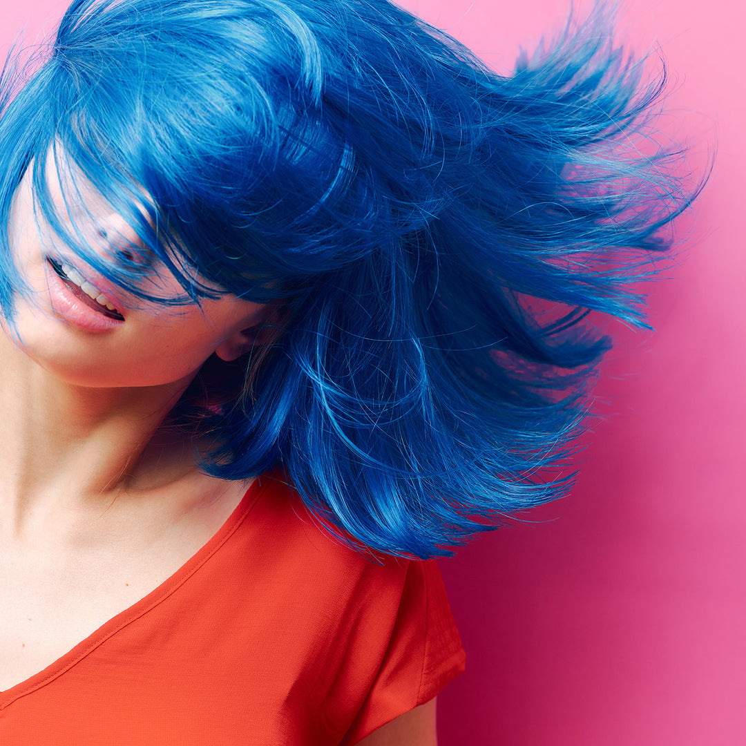 En person med levende blåt hår er i bevægelse, hvilket får deres hår til at vifte dramatisk ud. De er iført en orange skjorte og er sat mod en lyserød baggrund. Deres ansigt er delvist skjult af deres hår.