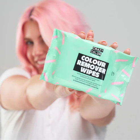 En person med pastelfarvet hår smiler og holder en turkis Crazy Color Color Remover Wipes/Farvefjerner-pakke op mod kameraet. Disse biologisk nedbrydelige klude fjerner skånsomt hårfarve; emballagen har pink og hvide detaljer på en lys baggrund.