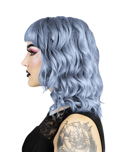 En person med lyseblåt krøllet hår og pandehår, opnået ved hjælp af Hermans Hårfarve Stella Blue (115 ml), bærer dramatisk makeup med mørk læbestift og øjenskygge. De er i en sort top med blondedetaljer og har en rosetatovering på armen mod en almindelig hvid baggrund.