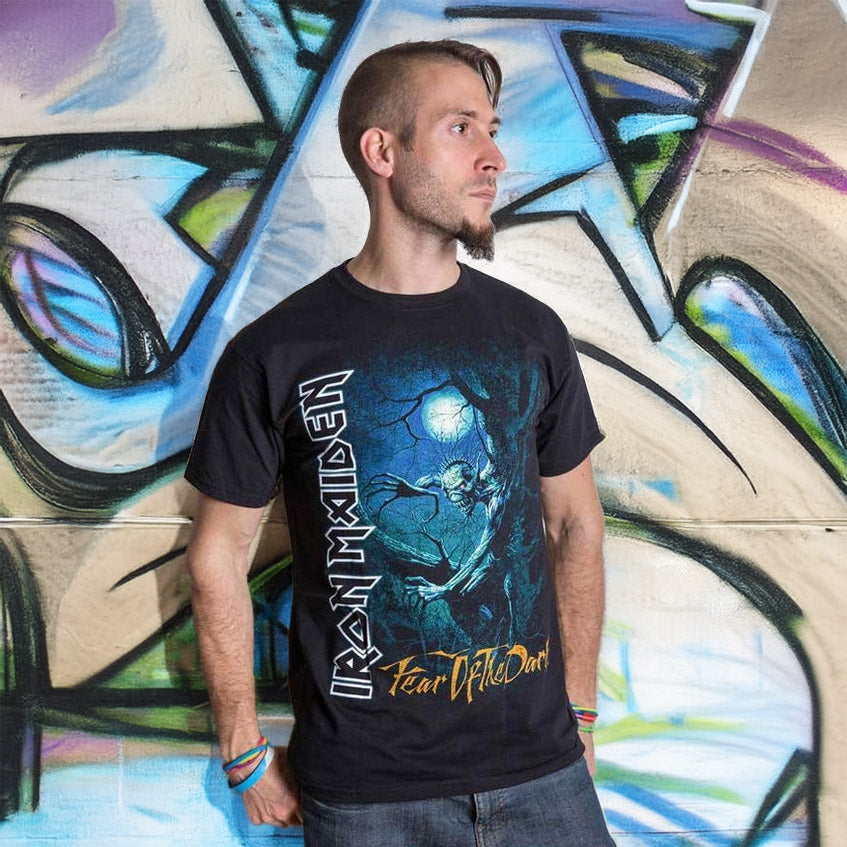 En mand med kort hår og skæg står op ad en farverig graffitivæg. Han er iført en sort Iron Maiden "Fear of the Dark" T-shirt og blå armbånd og kigger til siden med armene over kors.