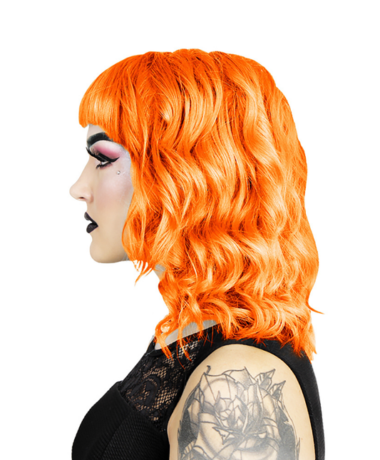 En person med lyst orange, bølget hår i bob-stil og pandehår fremviser Hermans Hårfarve Tara Tangerine (115 ml) af mærket Hermans. De bærer dramatisk makeup med sort læbestift og lyserød øjenskygge, og en tatovering er synlig på deres arm mod deres sorte blonde-detaljerede top, sat mod en hvid baggrund.