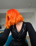 Indlæs billede i Gallery viewer, En person med Hermans Hårfarve Tara Tangerine lyst orange hår stylet i en bølget bob vender væk fra kameraet, iført en sort top og lædersele. I baggrunden er der et glimt af et køkkenområde i et hvidvægget rum.