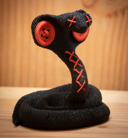 MM Figur Sacky Serpent (13 cm) er en sammenrullet sort figur med røde knapøjne og syede X-mærker, der står oprejst - ideel som hyldeudsmykning.